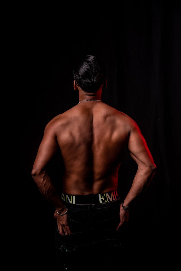 A shirtless man with a muscular build stands against a black background, viewed from behind.