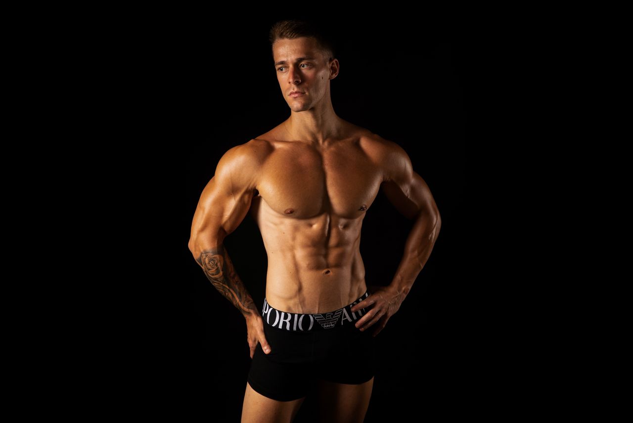 Shirtless male bodybuilder showcasing strong physique in a studio setting with a black background.