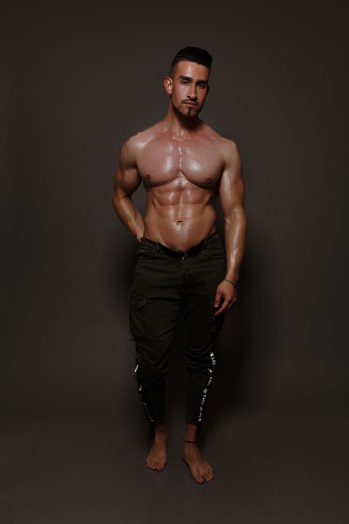 Full body shirtless male with dark hair and muscular torso standing against gray background with one hand in pocket